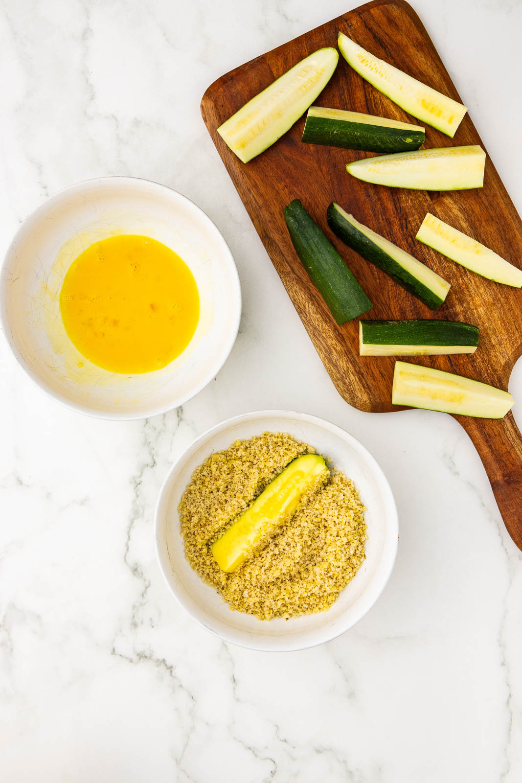 wooden cutting board with sliced zucchini, with two bowls containing beaten egg and panko mixture with zucchini