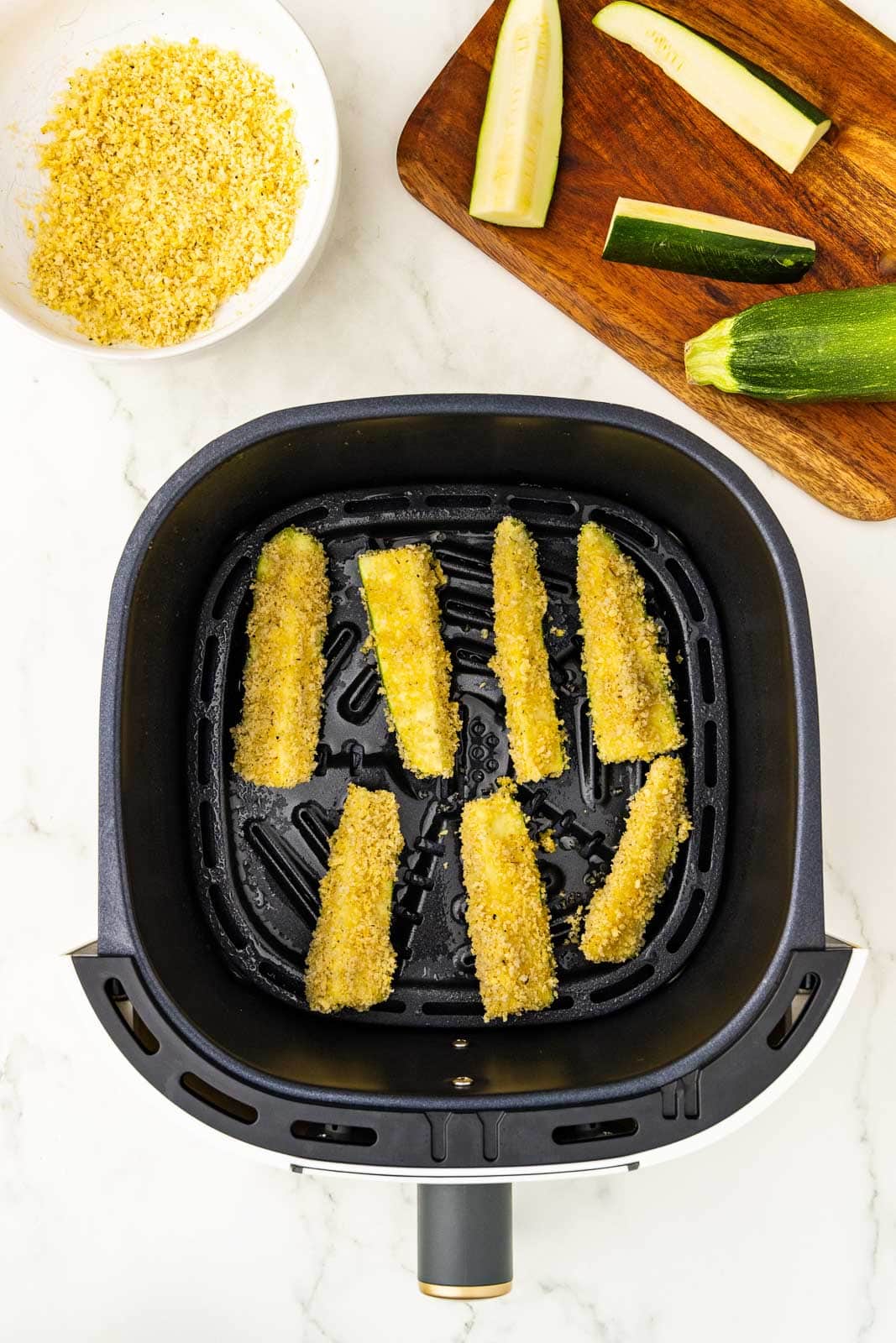 overhead view of air fryer containing zucchini covered in panko mixture, with small bowel of panko and wooden cutting board with zucchini in background