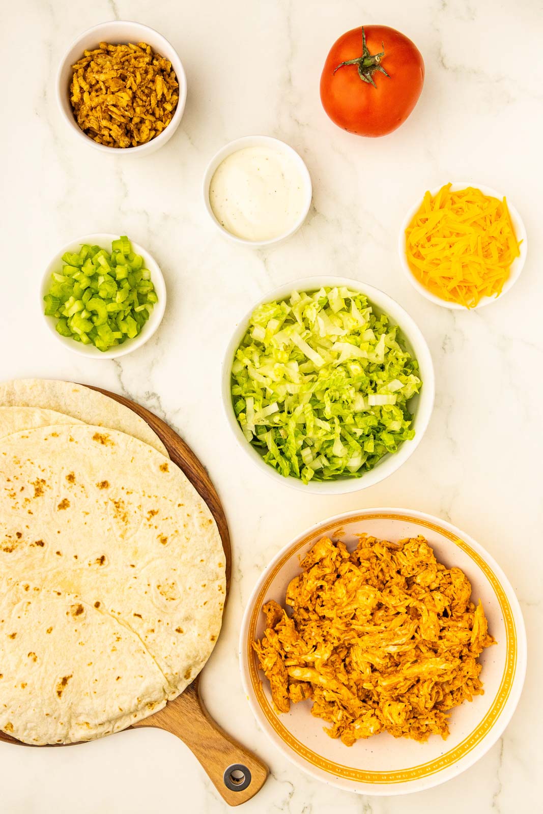 buffalo chicken wrap ingredients to include wooden cutting board with 4 flour tortillas, small white bowls containing leftover shredded buffalo chicken, shredded lettuce, shredded cheddar cheese, chopped celery, French fried onions, ranch dressing, and whole tomato