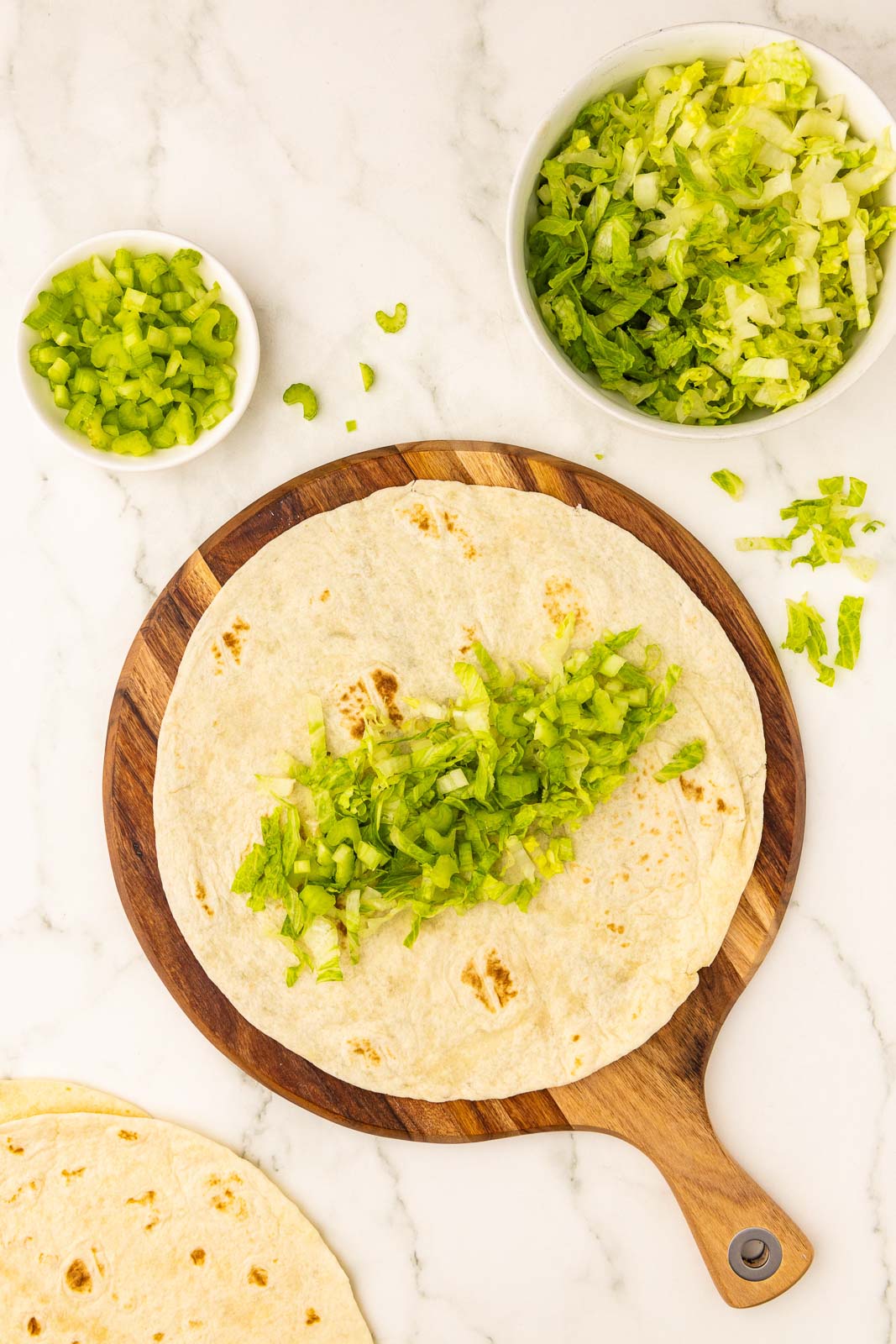 wooden cutting board with flour tortilla layered with chicken, lettuce and celery, surrounded by bowls of lettuce and celery, and tortillas on side