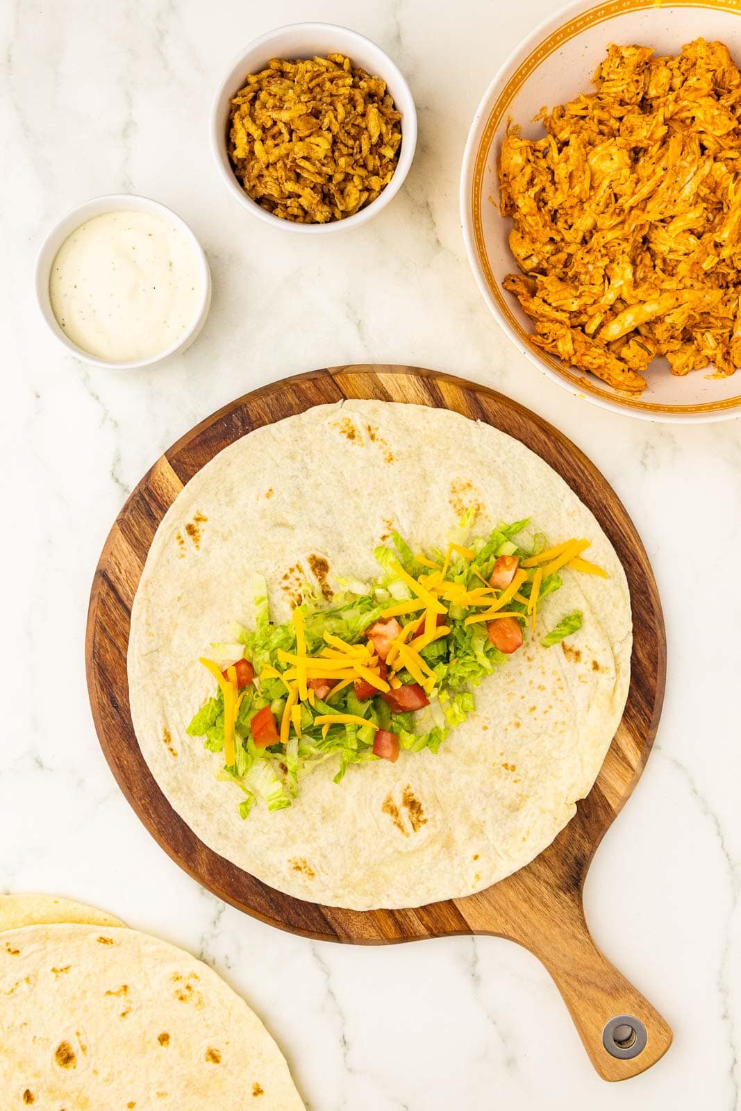 wooden cutting board with tortilla layered with chicken, lettuce, celery, tomato and cheese, with white bowls of chicken, fried onions, and ranch dressing in background, with tortillas on side