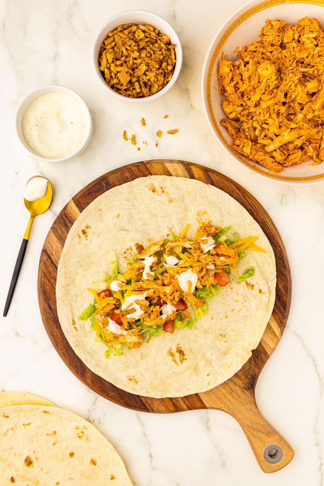 wooden cutting board containing tortilla layered with chicken, celery, lettuce, tomato, cheese and ranch surrounded by white bowls of chicken, fried onions, ranch dressing with gold tipped spoon and tortillas