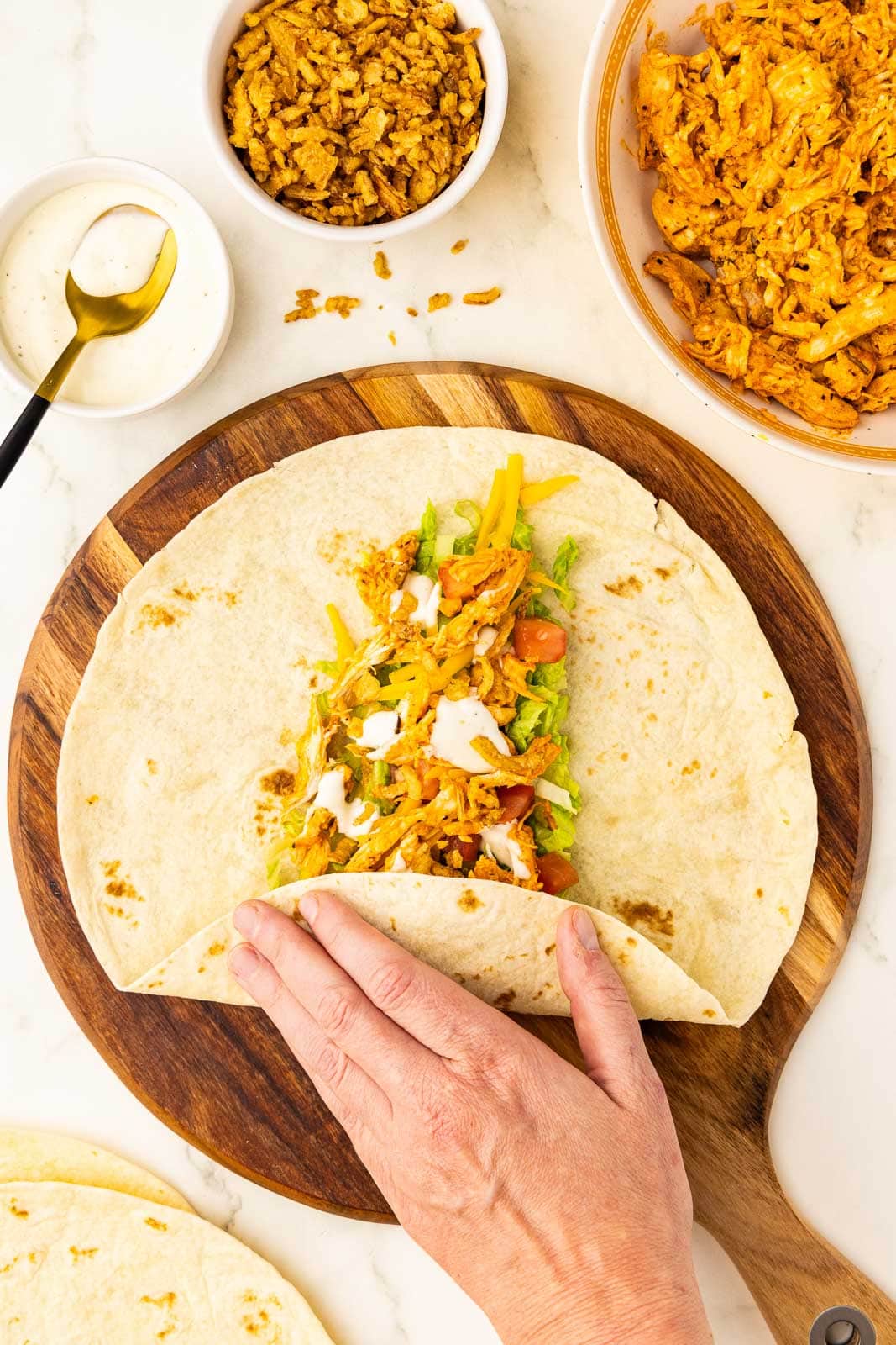 wooden cutting board with layered tortilla with hand folding tortilla with bowls of fried onions, chicken, ranch with gold tipped spoon, and tortillas in background
