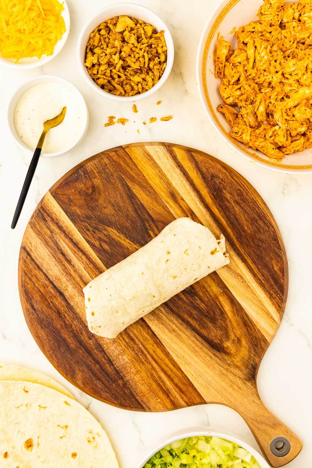 wooden cutting board with folded tortilla, with bowls of chicken, fried onions, cheese, ranch with gold tipped spoon, lettuce and tortillas in background