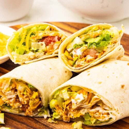 wooden cutting board with four halves of cut tortillas showing insides containing chicken, lettuce, celery, tomatoes, cheese, fried onions, with bowl of potato chips and fried onions