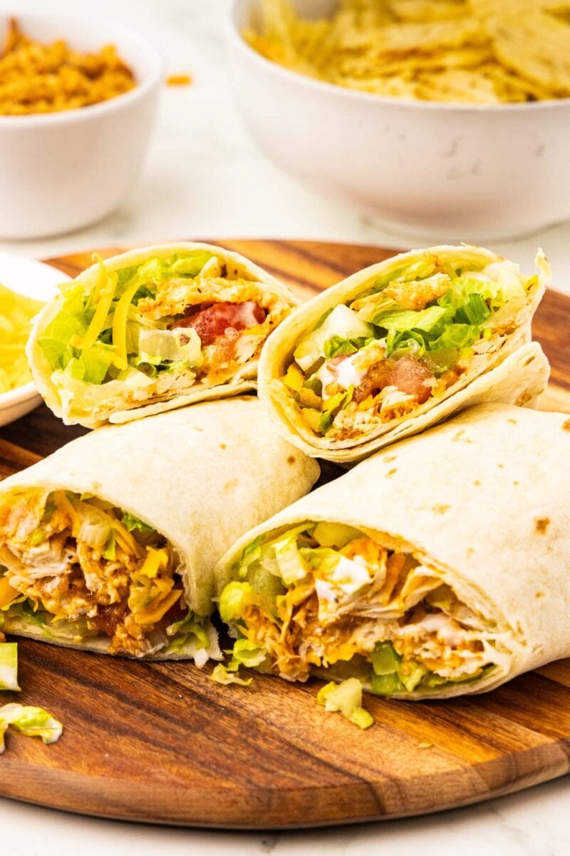 wooden cutting board with four halves of cut tortillas showing insides containing chicken, lettuce, celery, tomatoes, cheese, fried onions, with bowl of potato chips and fried onions