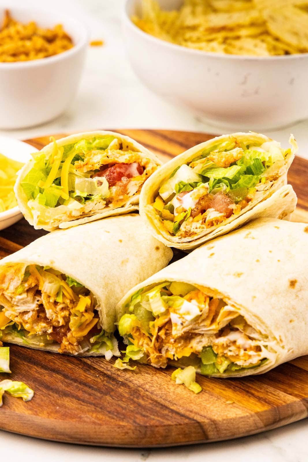 wooden cutting board with four halves of cut tortillas showing insides containing chicken, lettuce, celery, tomatoes, cheese, fried onions, with bowl of potato chips and fried onions
