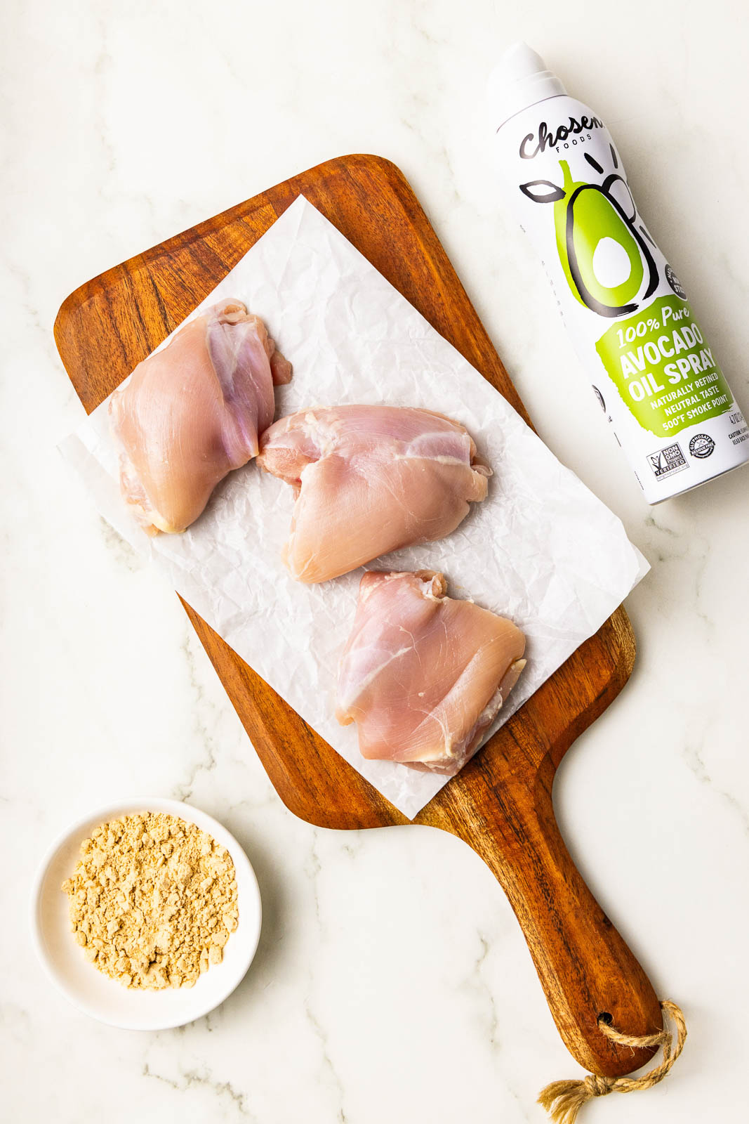 ingredients to include 3 chicken thighs on parchment paper lined wooden cutting board, surrounded by a can of avocado spray and white bowl containing seasoning