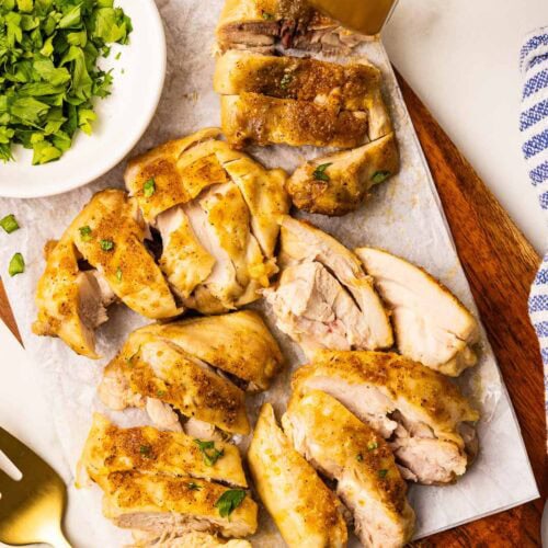 overhead view of sliced air fried chicken thighs on parchment lined wooden cutting board with white bowl of parsley, gold fork with blue and white striped dishcloth and air fryer in background