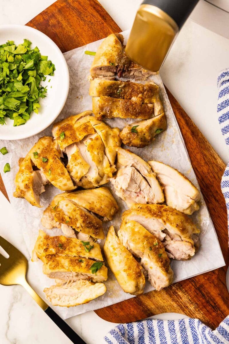 overhead view of sliced air fried chicken thighs on parchment lined wooden cutting board with white bowl of parsley, gold fork with blue and white striped dishcloth and air fryer in background
