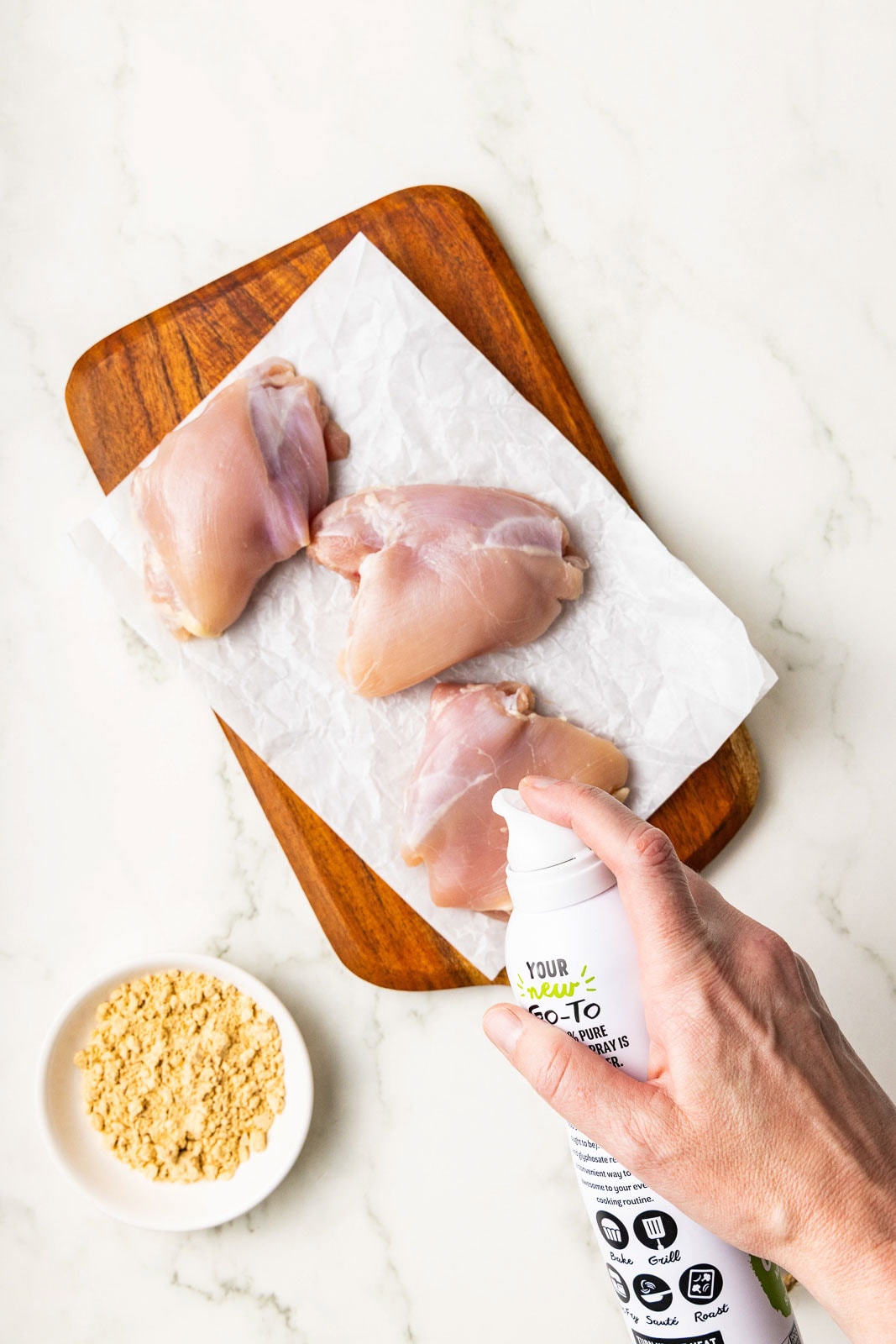 3 chicken thighs on parchment paper lined wooden cutting board, with person spraying avocado spray on thighs and white bowl containing seasoning