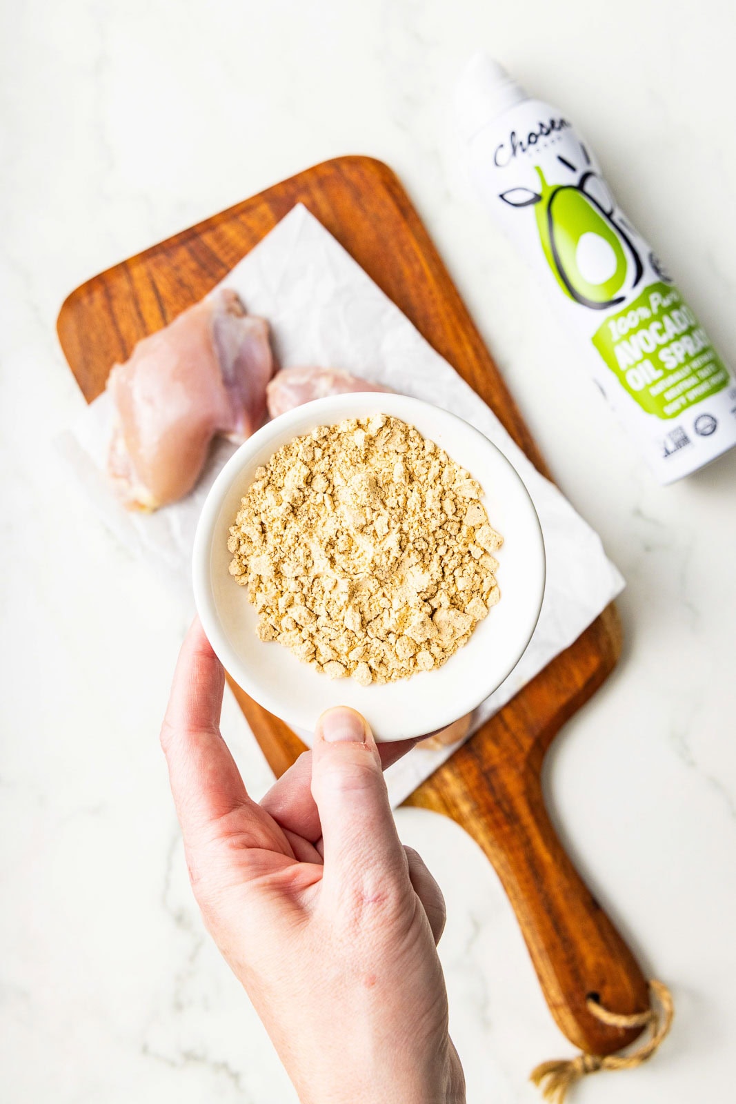 closeup of white bowl with seasoning with 3 chicken thighs on parchment paper lined wooden cutting board and a can of avocado spray