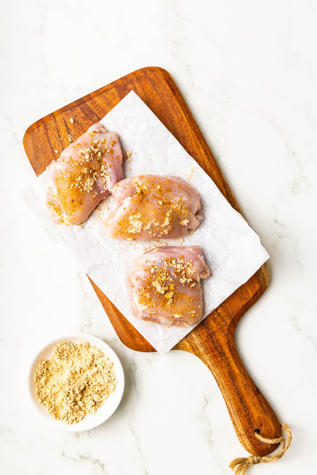 3 seasoned chicken thighs on parchment lined wooden cutting board with white bowl containing seasoning on side