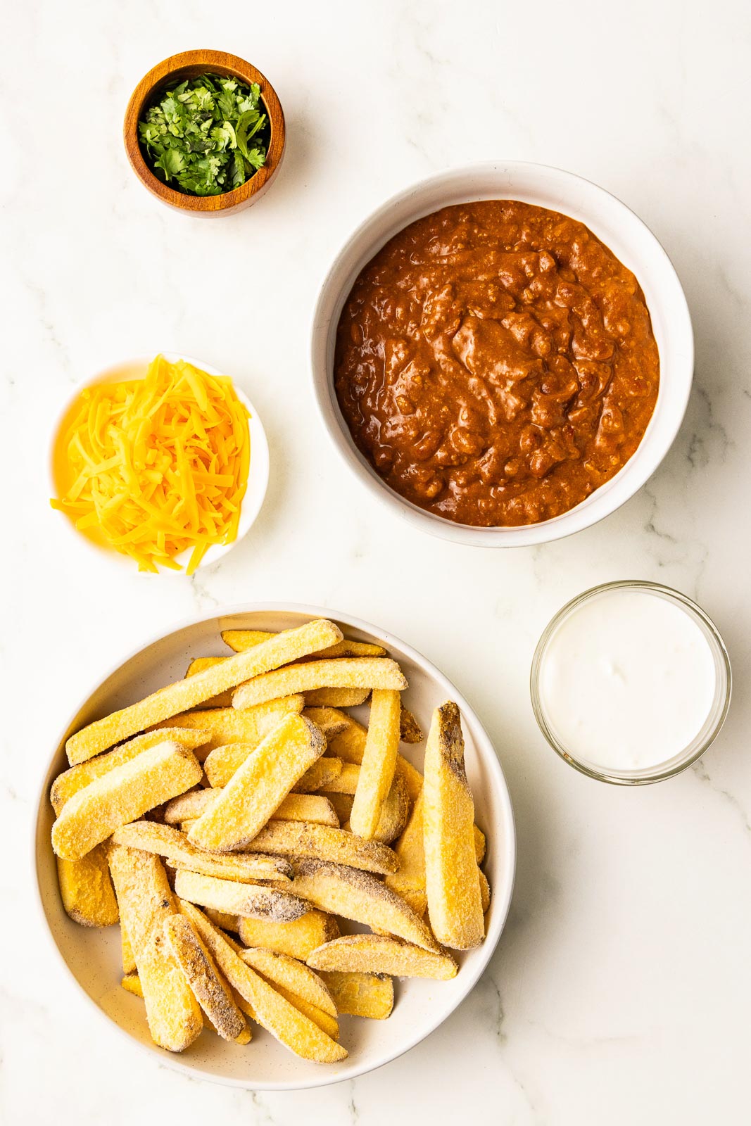 ingredients in bowls with frozen french fries, chili, shredded cheddar cheese, sour cream and chopped cilantro
