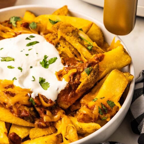 side view of bowl of French fries with chili, cheddar cheese, sour cream and cilantro in front of a white air fryer with gold handle with black and white checked dish towel on side