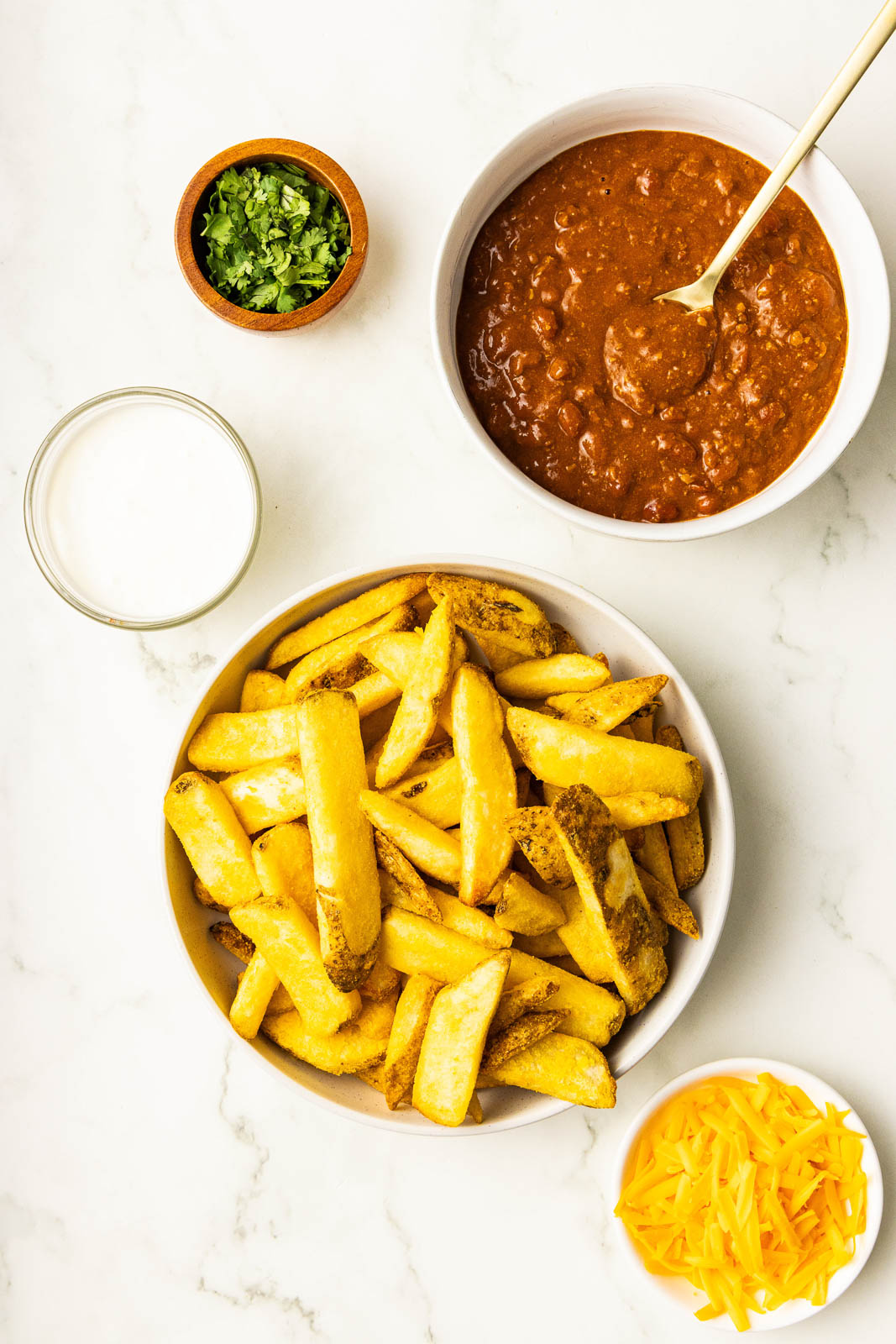 white bowls of air fried french fries, chili with spoon, shredded cheese, sour cream and small wooden bowl with chopped cilantro
