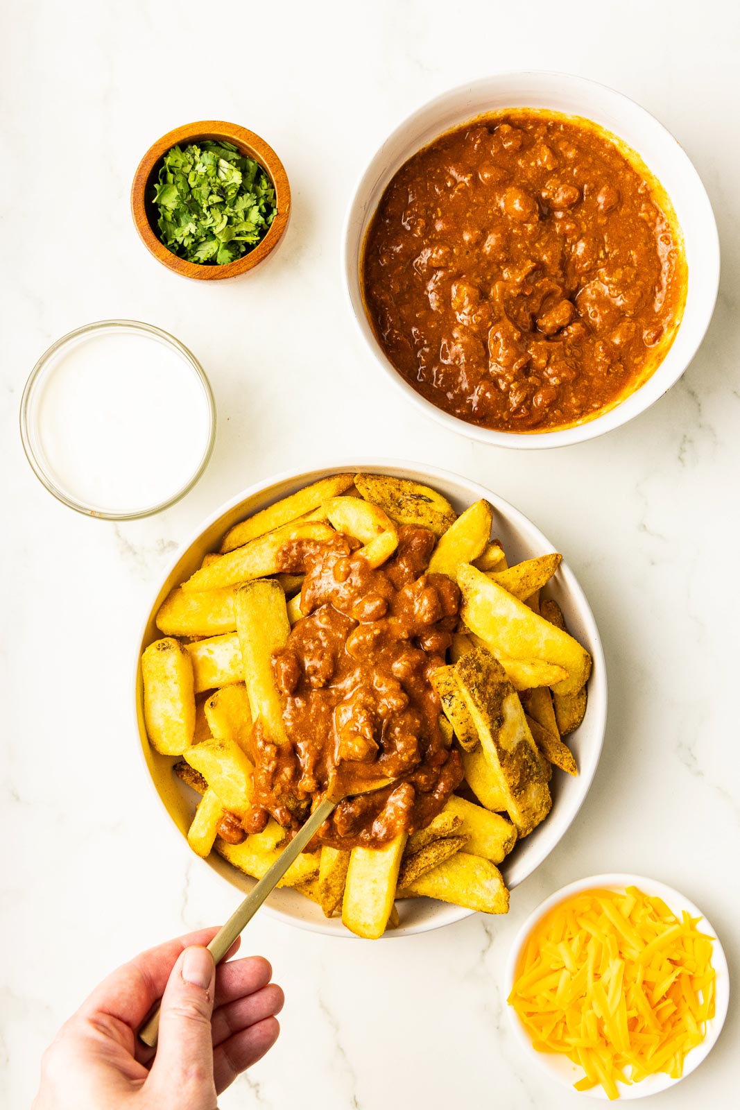 overhead view of bowl of air fried french fries with chili being added with gold spoon, surrounded by bowls of shredded cheddar cheese, sour cream, chili and small wooden bowl of chopped cilantro