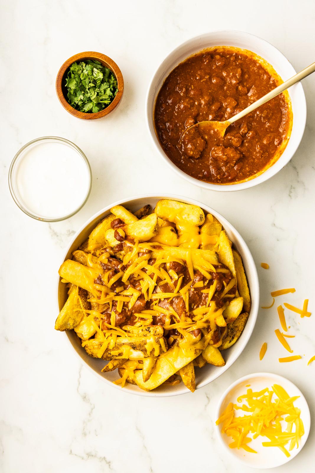 overhead view of bowl of air fried french fries with chili and cheese added, surrounded by bowls of half-empty shredded cheddar cheese, sour cream, chili with gold spoon, and small wooden bowl of chopped cilantro