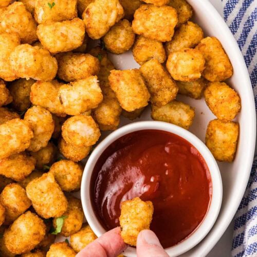 white plate filled with air fried tater tots with hand dipping tater tot into bowl of ketchup, with blue and white dishcloth on side
