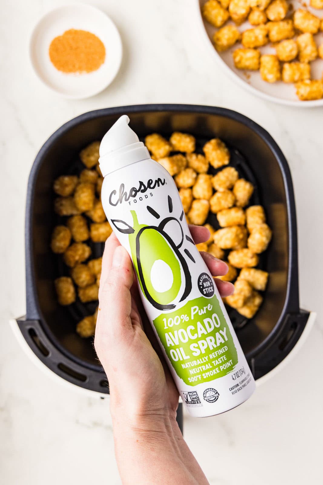 closeup of avocado spray with air fryer filled with tater tots, white bowl of Cajun seasoning and white bowl of tater tots in background