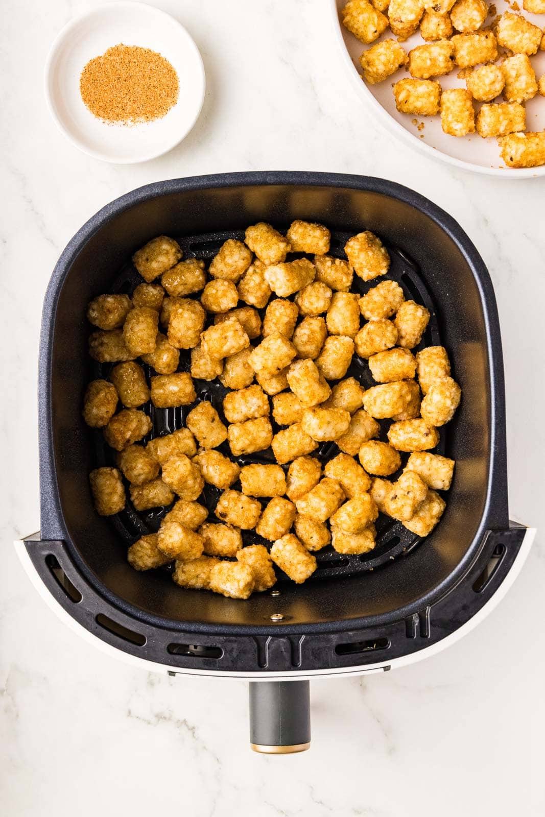 black air fryer filled with tater tots surrounded by white bowl of Cajun seasoning and white bowl of tater tots