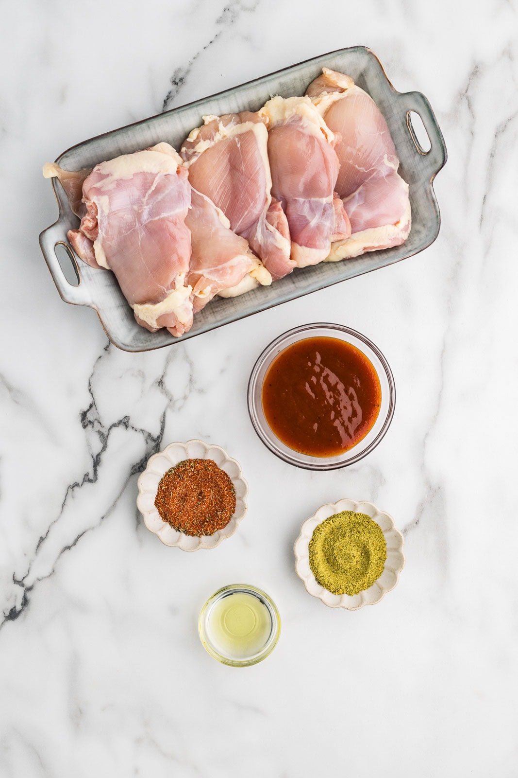 container of chicken thighs with bowls of BBQ sauce, olive oil, garlic and herb seasoning, and Cajun seasoning