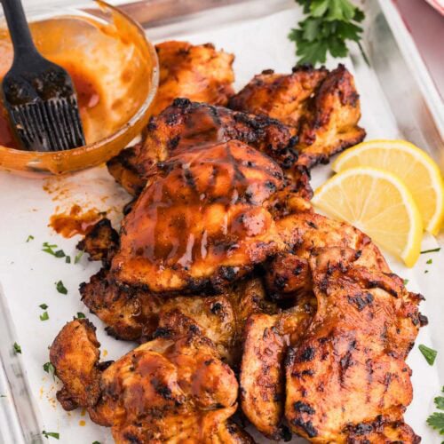 overhead view of grilled bbq chicken thighs on parchment lined baking sheet, with lemon slices, parsley sprigs and bowl of BBQ sauce and brush on side, with silver tongs in background