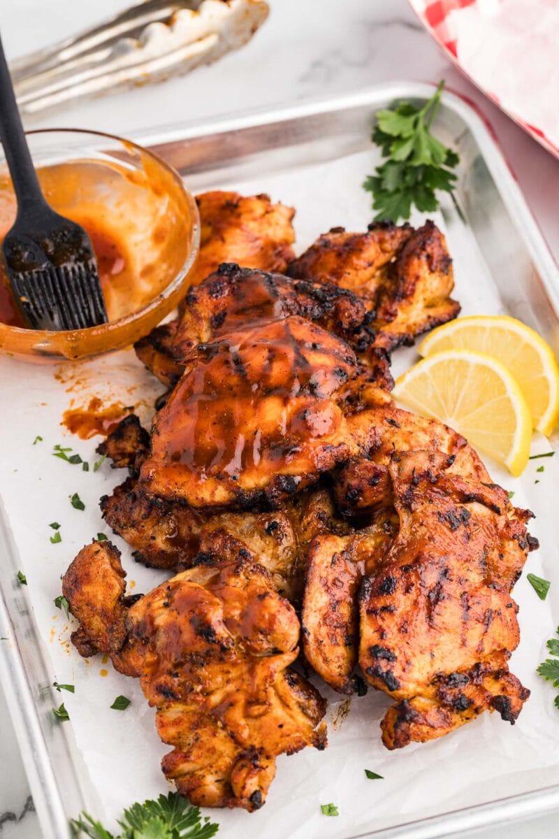 overhead view of grilled bbq chicken thighs on parchment lined baking sheet, with lemon slices, parsley sprigs and bowl of BBQ sauce and brush on side, with silver tongs in background