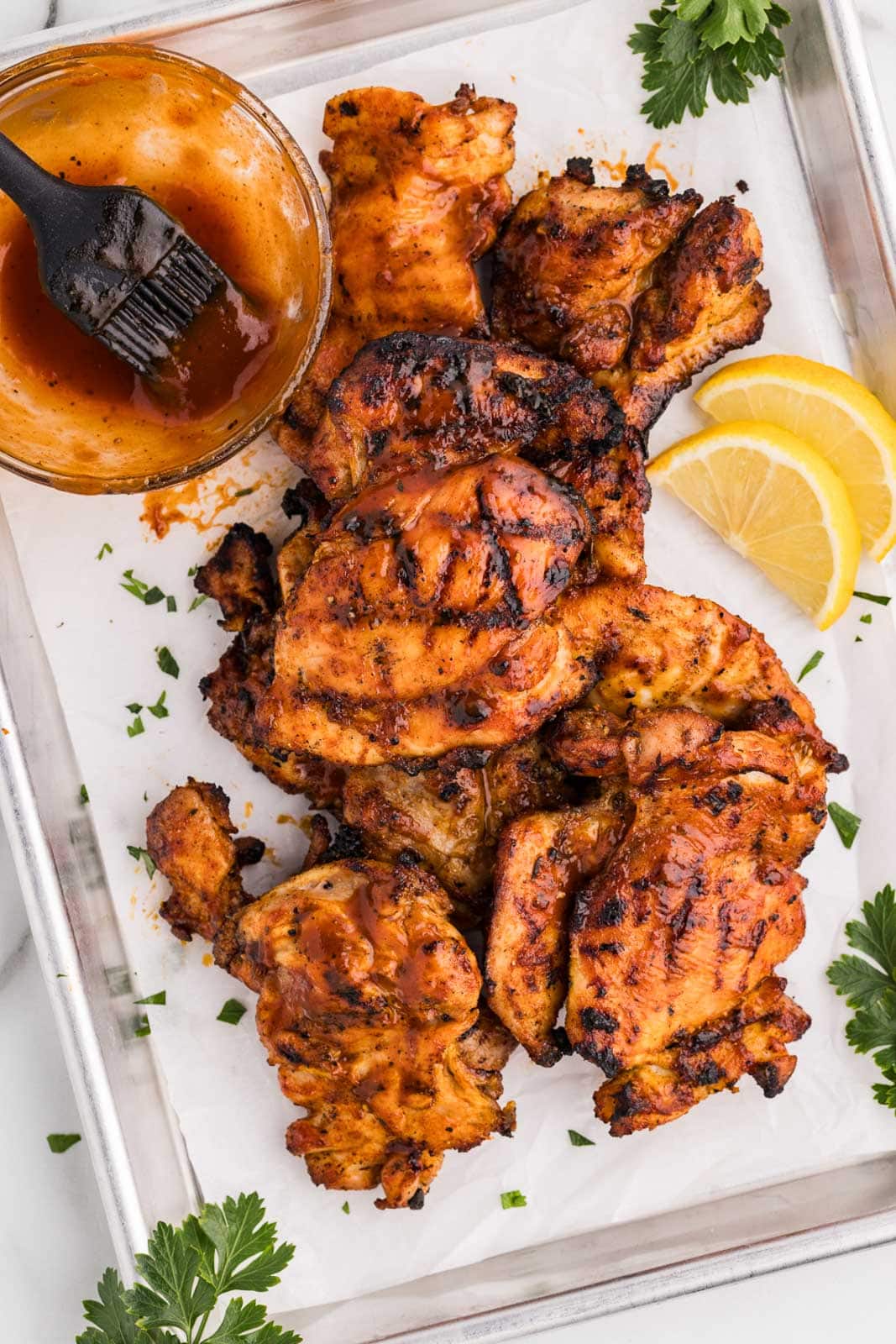 parchment lined baking sheet with grilled chicken thighs brushed with BBQ sauce, with lemon slices, parsley sprigs and bowl of BBQ sauce and brush on side