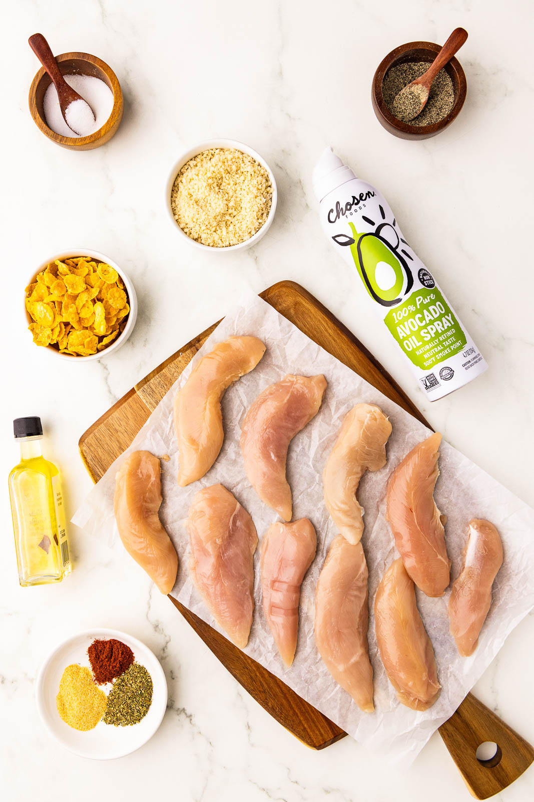 ingredients to include 10 chicken tenders on parchment lined wood cutting board, surrounded by bowls of salt, pepper, cornflakes, panko, oil, avocado oil spray, spices