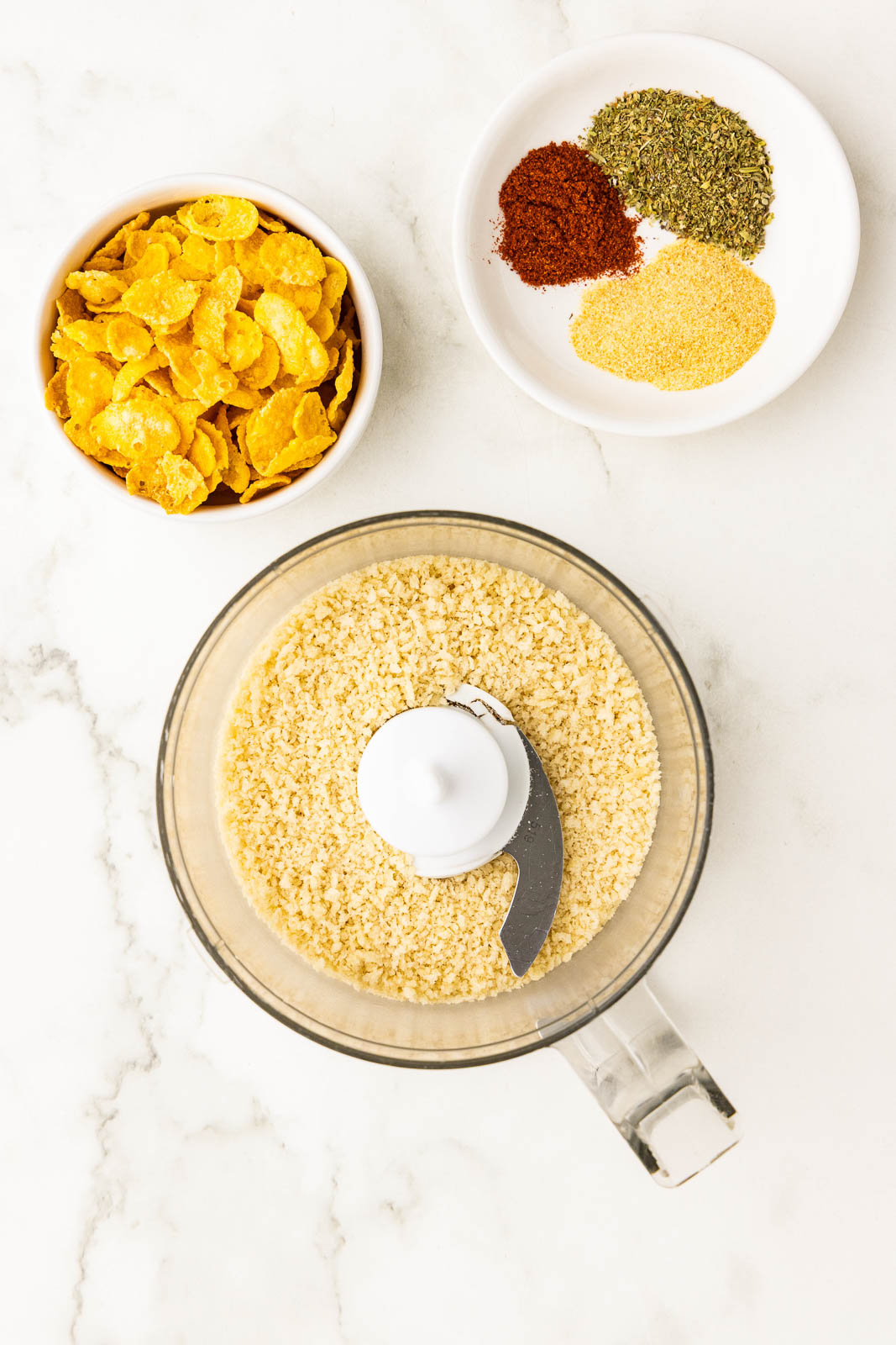 overhead view of food processor containing panko, surrounded by 2 white bowls of cornflakes and spices