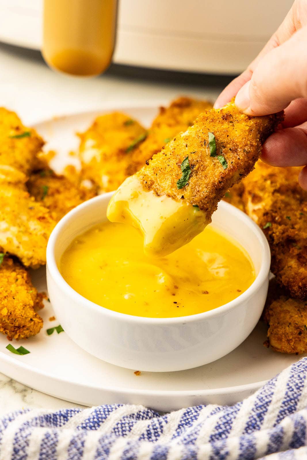 closeup of one chicken tenders being dipped in mustard sauce with white plate of chicken tenders, blue and white dishcloth, and air fryer in background