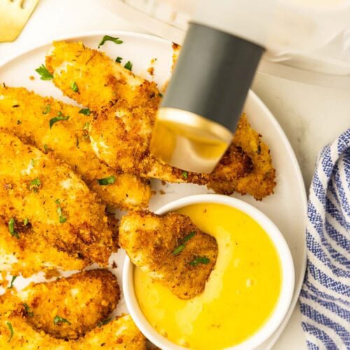 overhead view of chicken tenders on white plate with white bowl of mustard sauce containing a chicken tenders, with blue and white dishcloth, gold fork and air fryer in the background