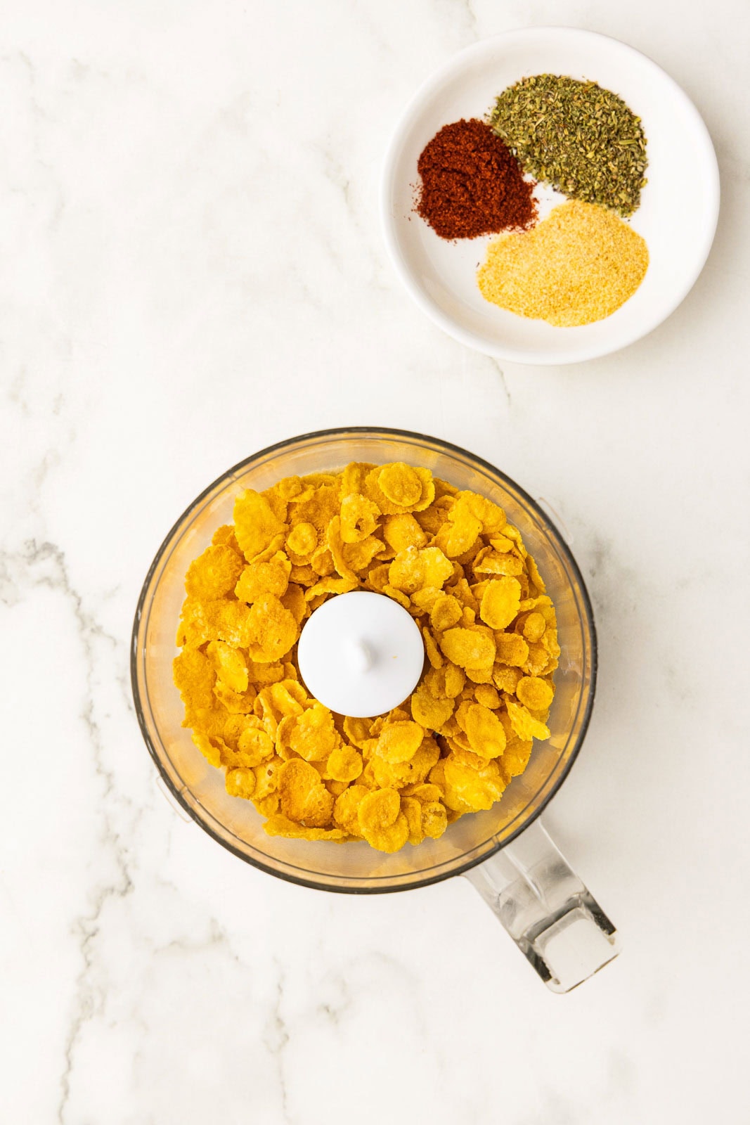 overhead view of food processor containing panko and cornflakes, surrounded by white bowl of spices