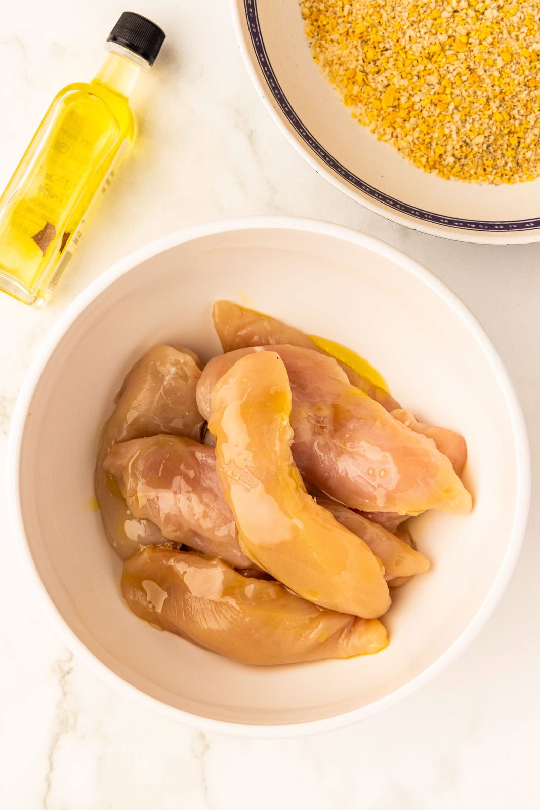 white bowl containing chicken tenders covered by oil, with another bowl containing processed mixture