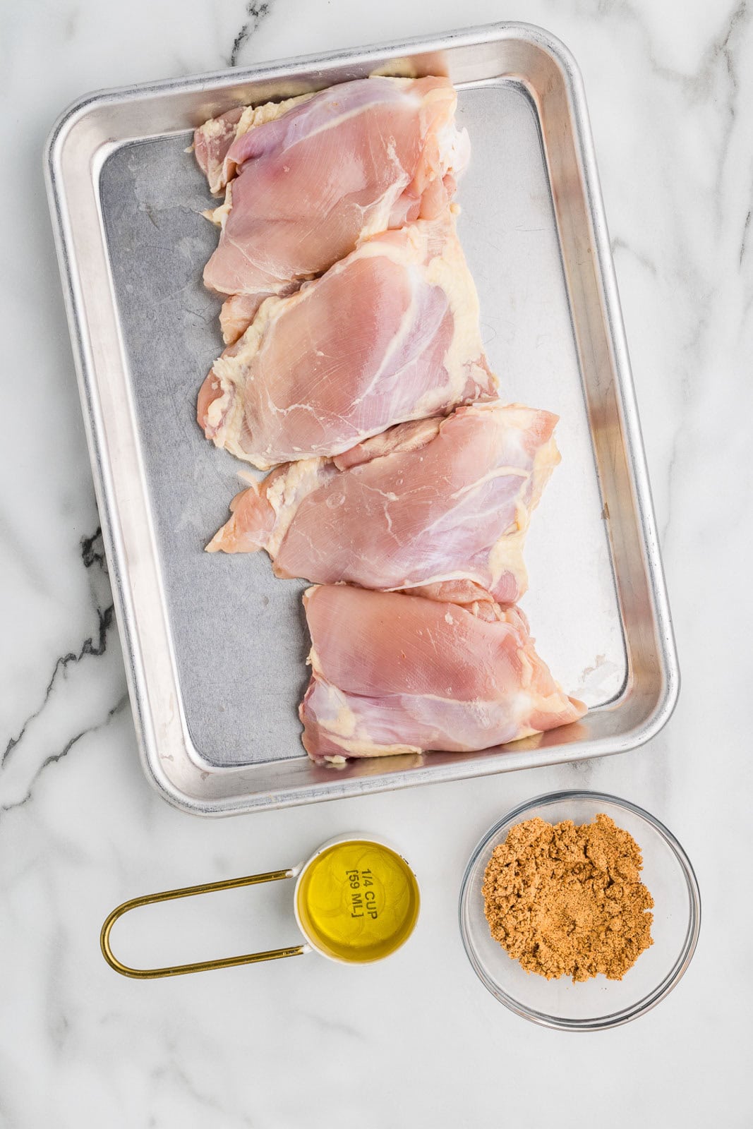 on cookie sheet ingredients including 4 chicken thighs with measuring spoon of olive oil and small clear bowl of seasoning