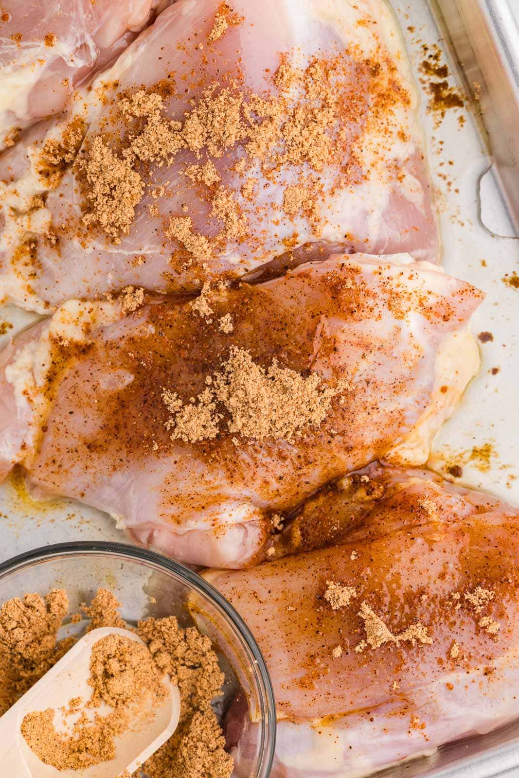 overhead view of chicken thighs on baking sheet, that have been oiled and seasoned, with small glass bowl of seasoning on side