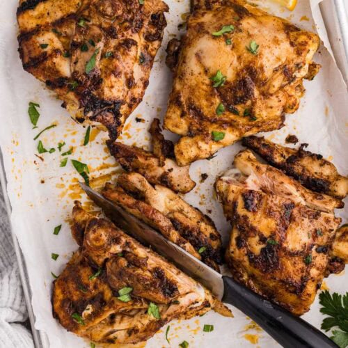 parchment covered baking sheet with 4 chicken thighs, with one being sliced by black handled knife, with lemon slices on side