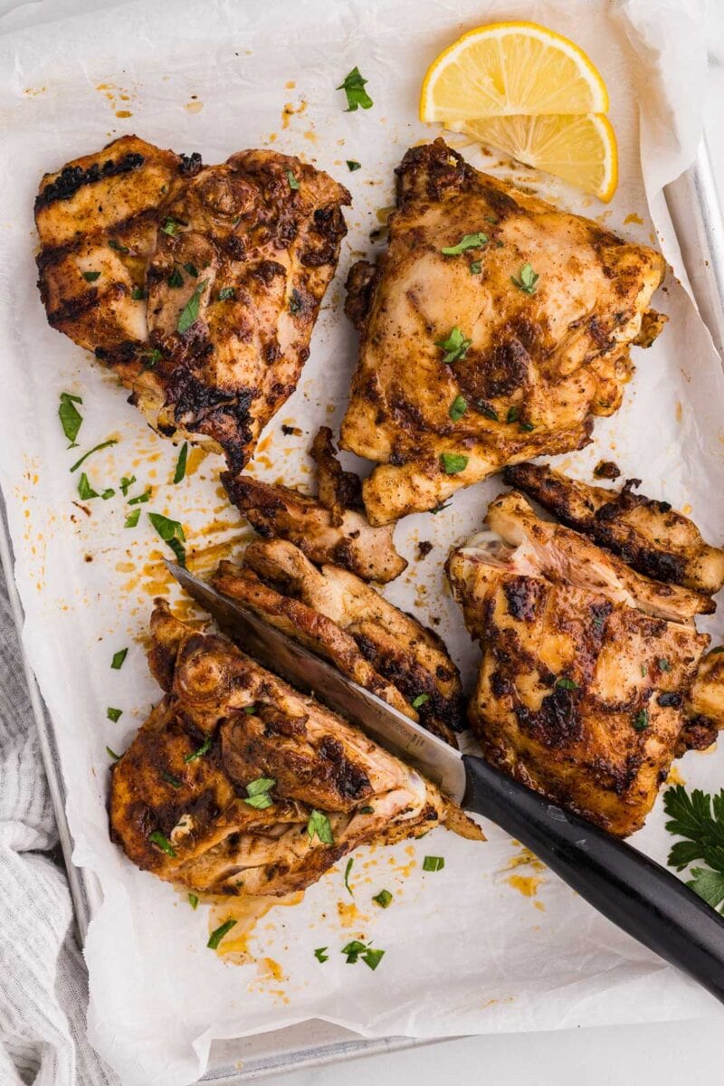 parchment covered baking sheet with 4 chicken thighs, with one being sliced by black handled knife, with lemon slices on side