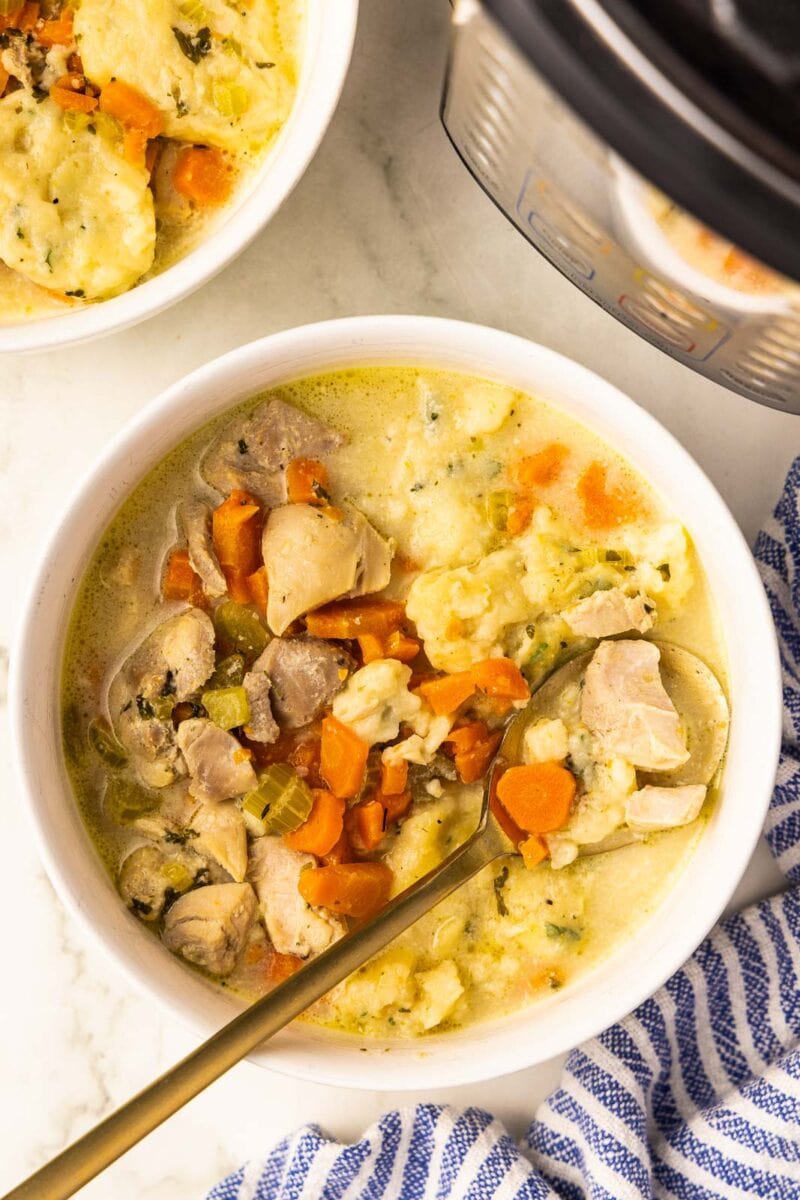 white bowl of chicken and dumplings with gold spoon, another bowl of chicken and dumplings, instant pot and blue and white striped dish towel in background