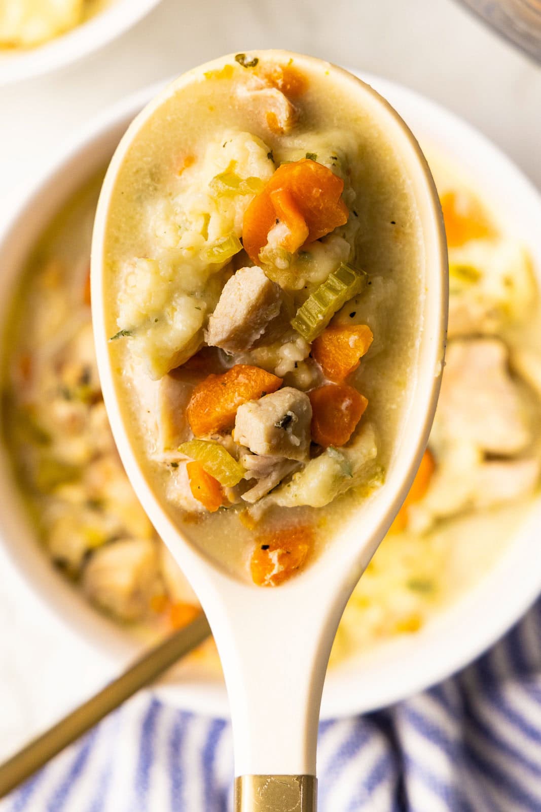 closeup of chicken and dumplings in large white spoon, over bowl of chicken and dumplings with gold spoon in background