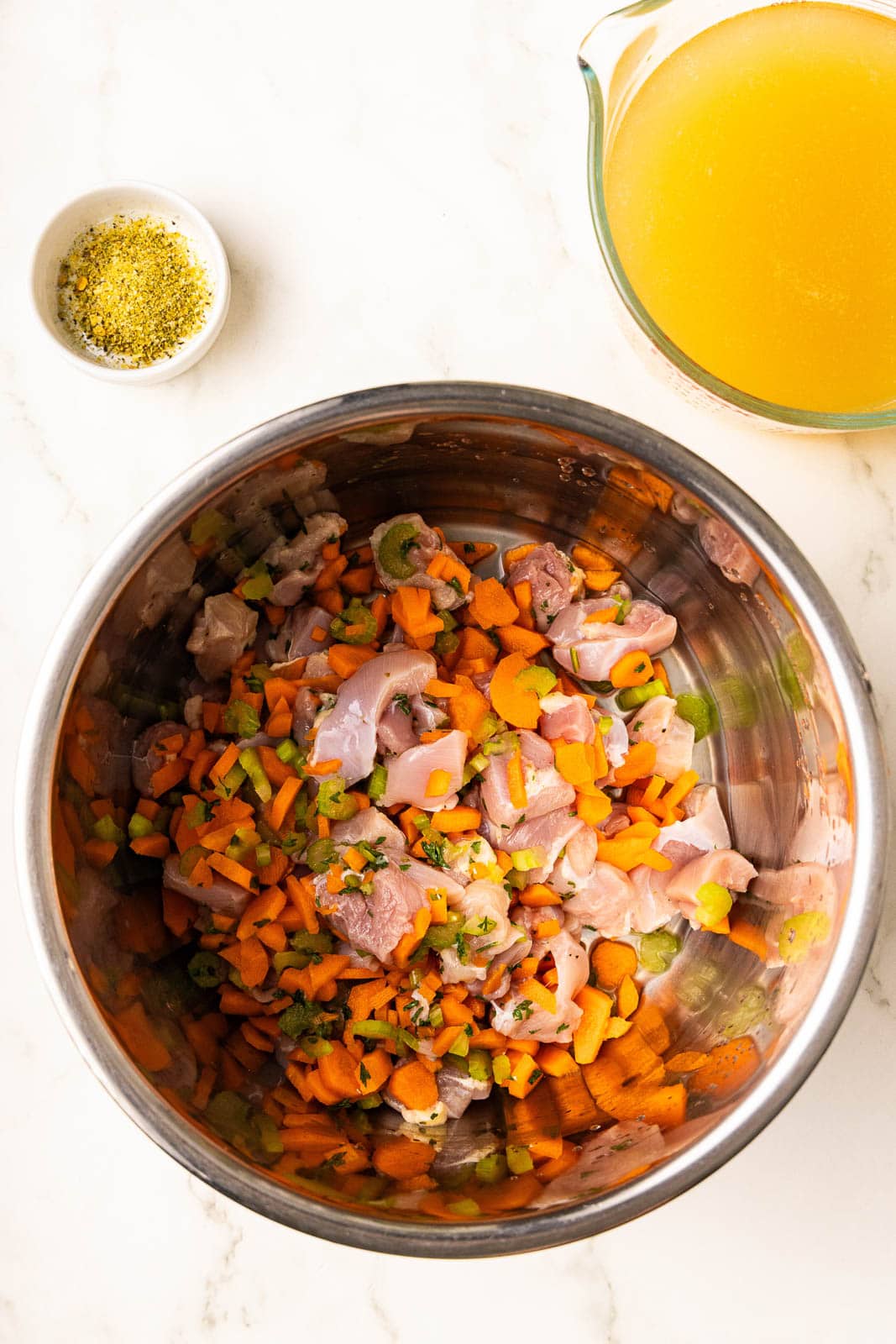 overhead view of instant pot containing diced chicken, carrots and celery, with bowl of seasoning and glass measuring cup of chicken broth