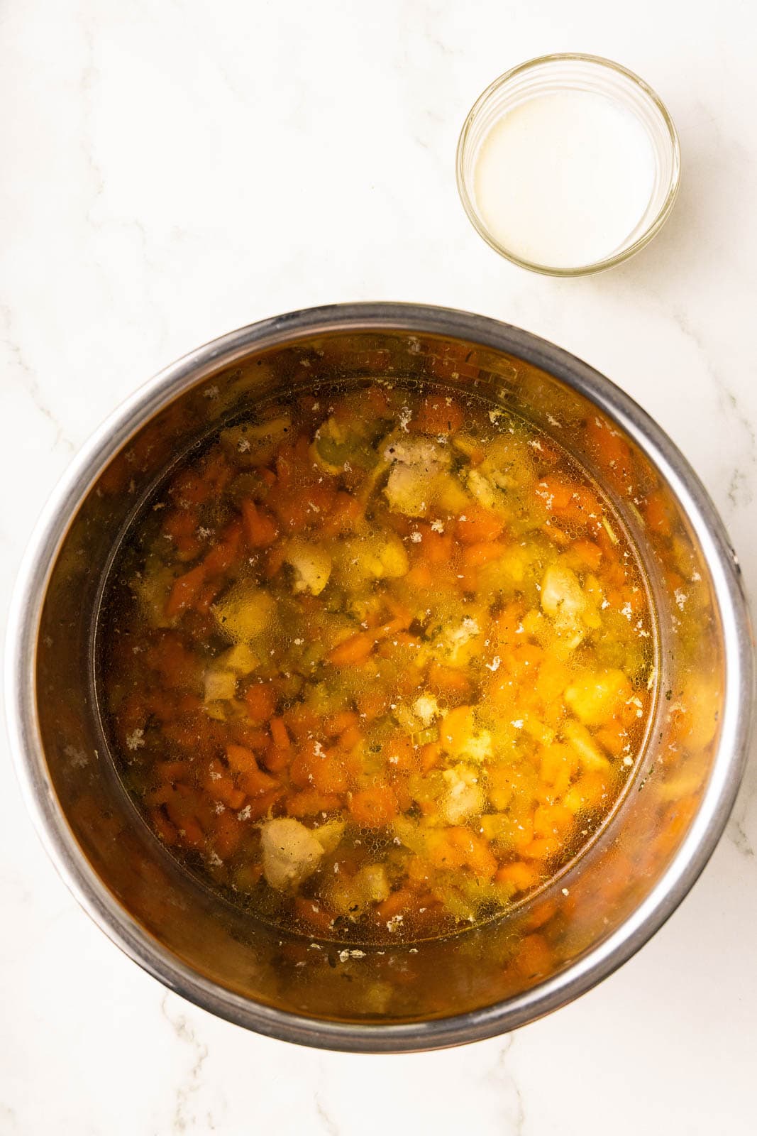 overhead view of instant pot containing diced chicken, carrots and celery, seasoning and chicken broth, with container of cornstarch and cream mixture on side