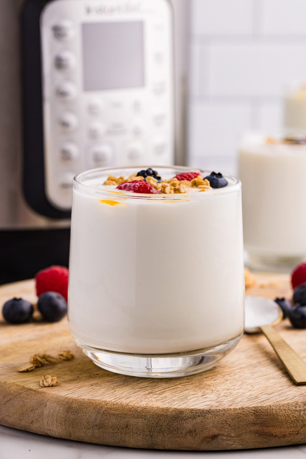 closeup of glass on top of wood circular cutting board, containing yogurt with blueberries, raspberries and granola on top, with scattered fruit on side with gold spoon and instant pot and another glass of yogurt in background