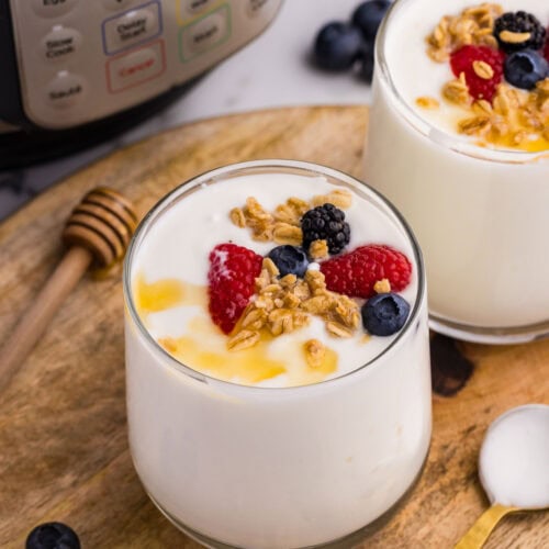 overhead view of 2 glasses of yogurt with fruit and drizzled with honey on round wooden cutting board, with gold spoon, scattered fruit and honey dipper on side, with instant pot and fruit in background