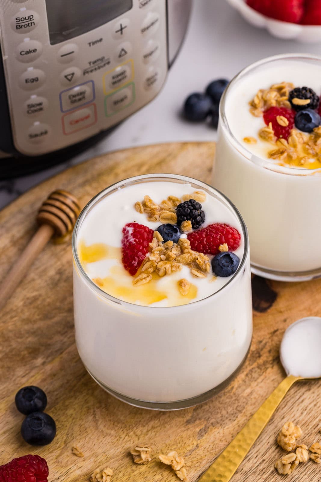 overhead view of 2 glasses of yogurt with fruit and drizzled with honey on round wooden cutting board, with gold spoon, scattered fruit and honey dipper on side, with instant pot and fruit in background