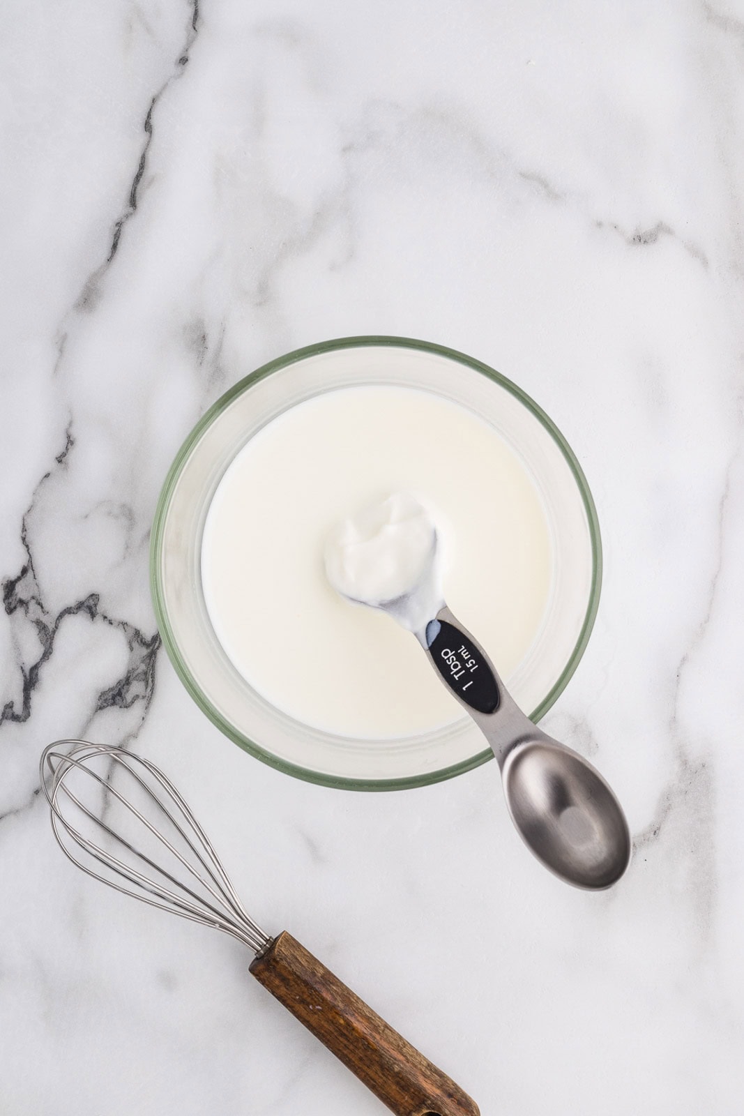 clear bowl with milk and measuring spoons with yogurt and whisk with wooden handle on side