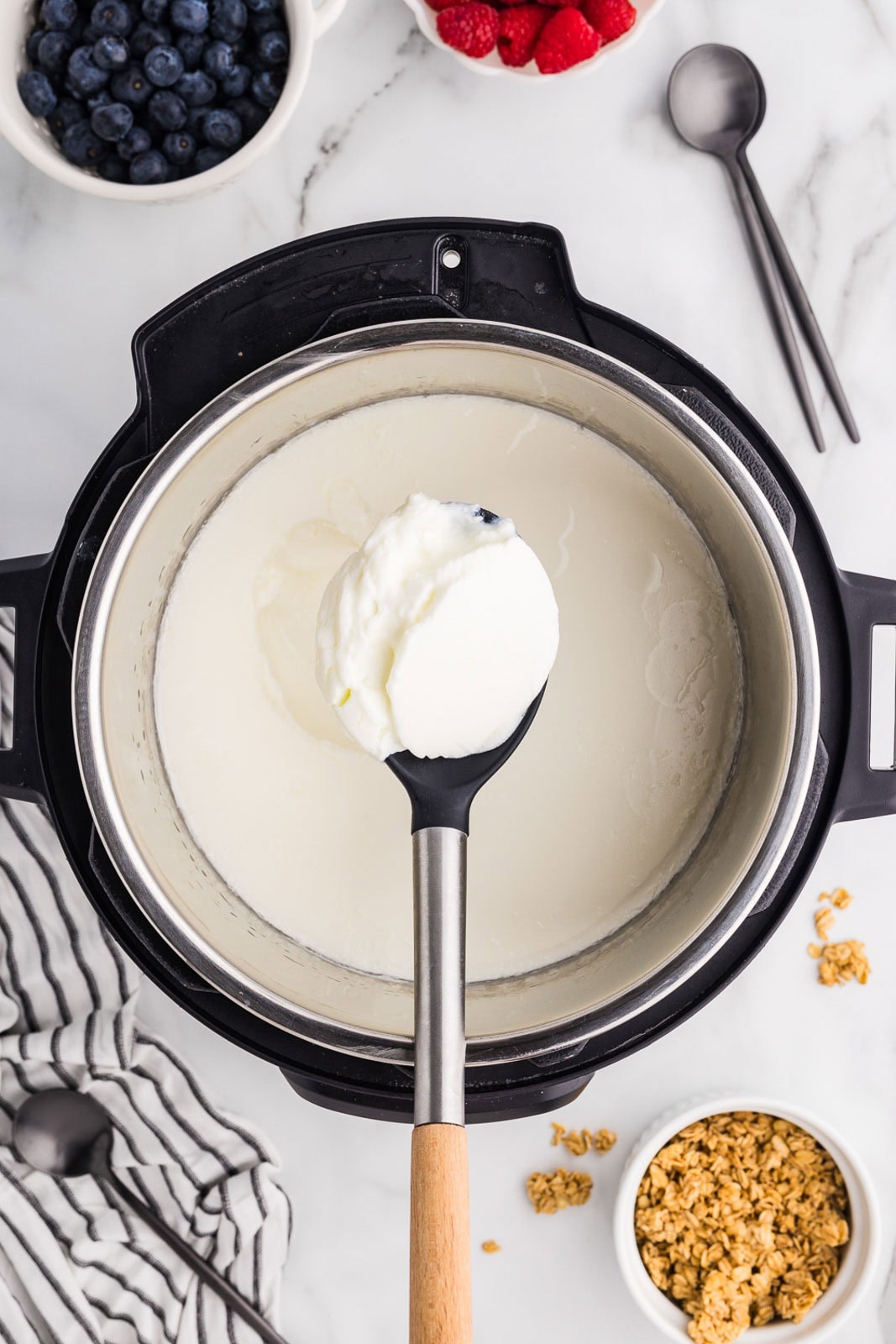 overhead view of instant pot containing yogurt with large wooden handled spoon containing yogurt, surrounded by small bowls of blueberries, raspberries, granola, with small spoons and a striped dish towel