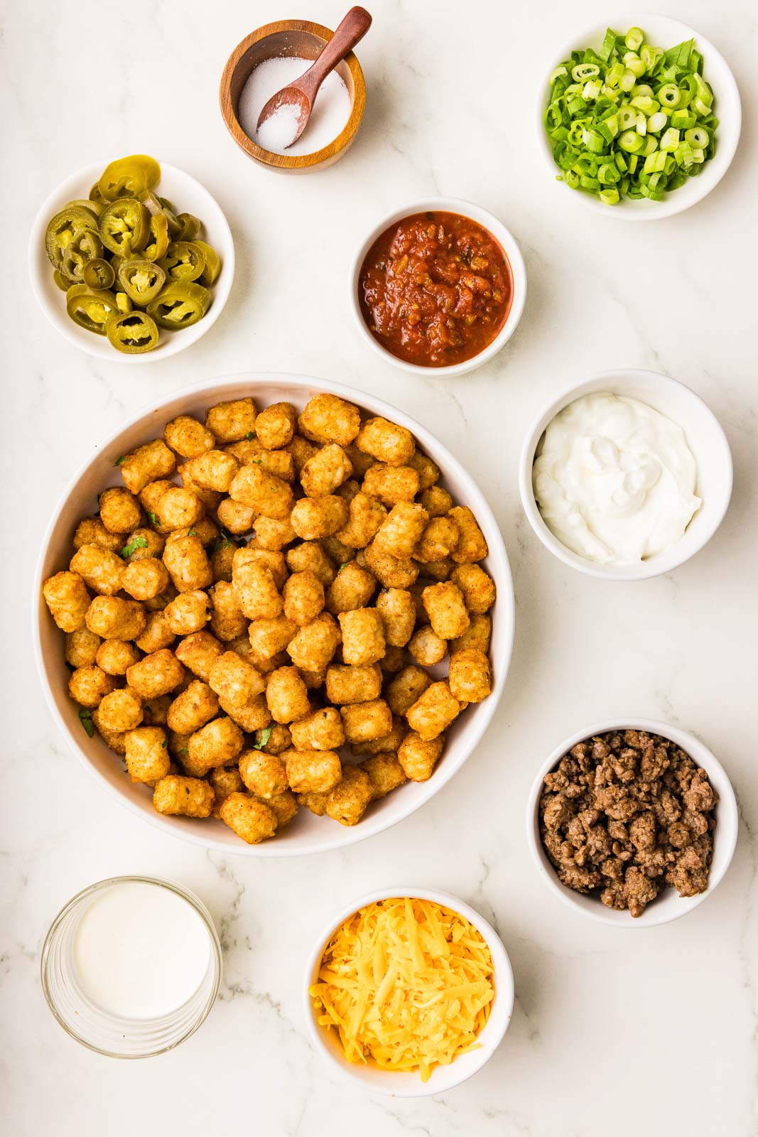 ingredients including large white bowl of tater tots surrounded by small bowls of taco-seasoned ground beef, shredded cheddar cheese, sour cream, salsa, green onion, sliced jalapeno peppers, salt and heavy cream