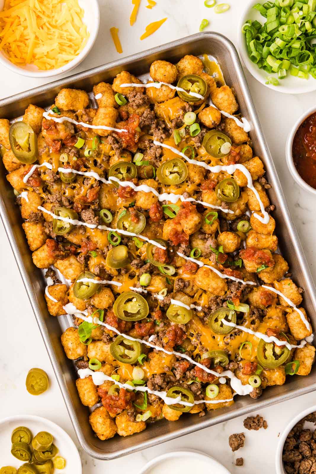 overhead view of completed tater tots on sheet pan drizzled with sour cream mixture, with bowls of cheddar cheese, green onions, jalapeno, salsa, ground beef and sour cream mixture